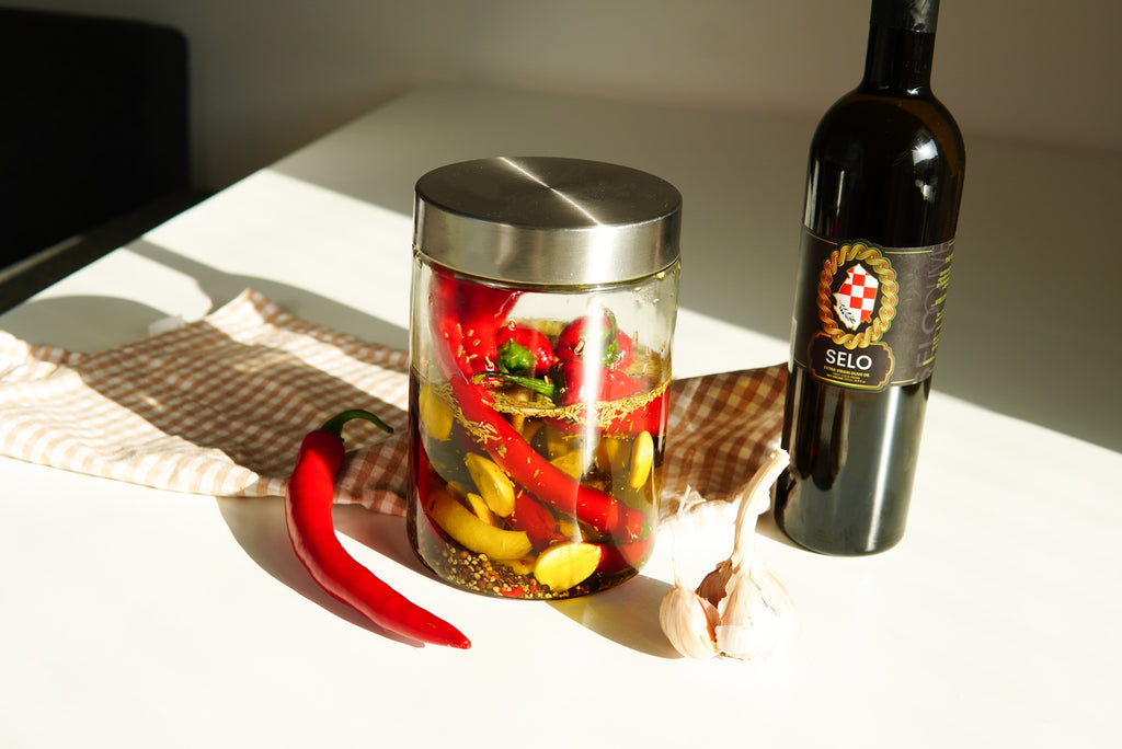 A sunlit jar of pickled chilies, garlic, rustic cloth, and Selo Croatian Olive Oil bottle on a white table.