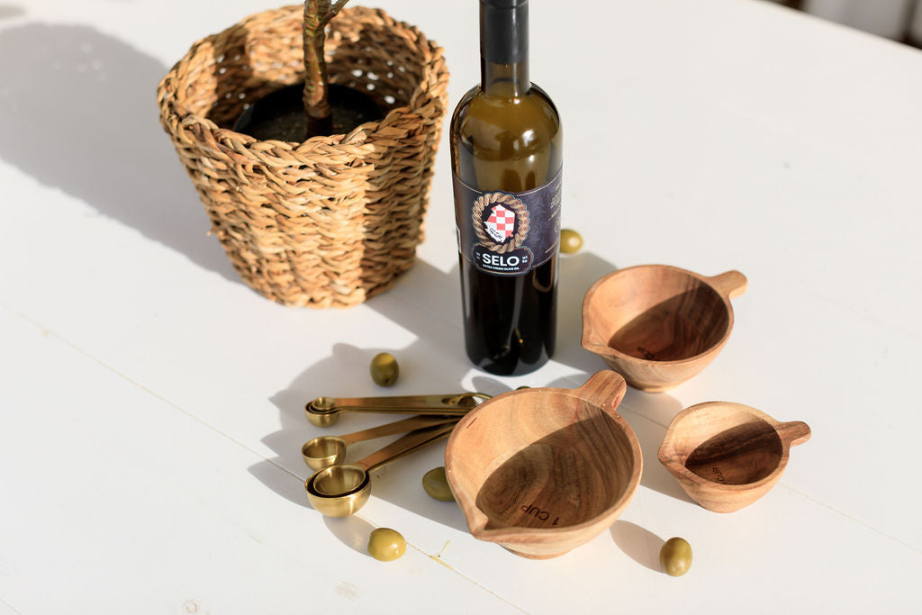 Selo olive oil bottle with measuring spoons and wooden bowls on white surface with wicker plant basket.