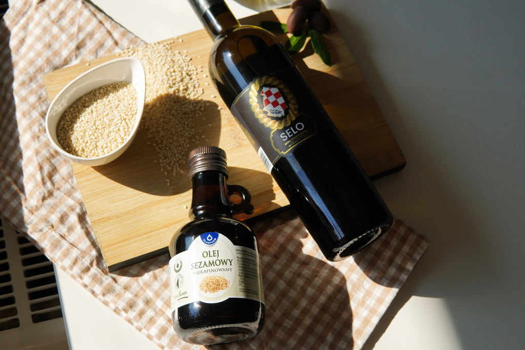 Resting on a draped tablecloth atop a pristine marble countertop, two elegant black bottles stand side by side: one of sesame oil, the other of olive oil. Nearby, a bowl brimming with whole sesame seeds offers a nod to the oil's origins.